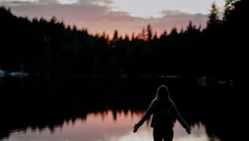 woman standing with hands spread on side facing lake at sundown