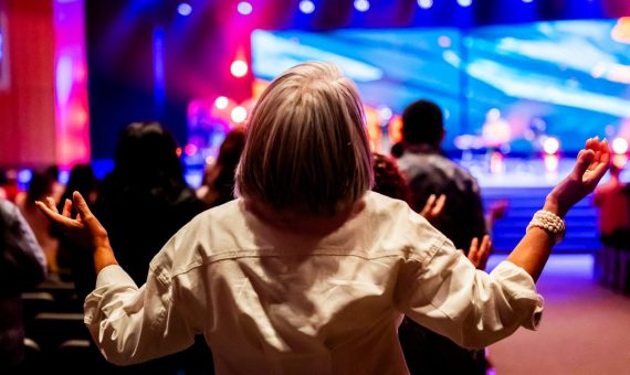 A vibrant indoor worship service with people raising hands in praise and colorful stage lighting.