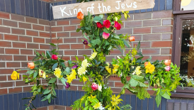 A cross adorned with flowers and greenery. There is a sign at the top saying 'King of the Jews'.