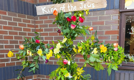 A cross adorned with flowers and greenery. There is a sign at the top saying 'King of the Jews'.