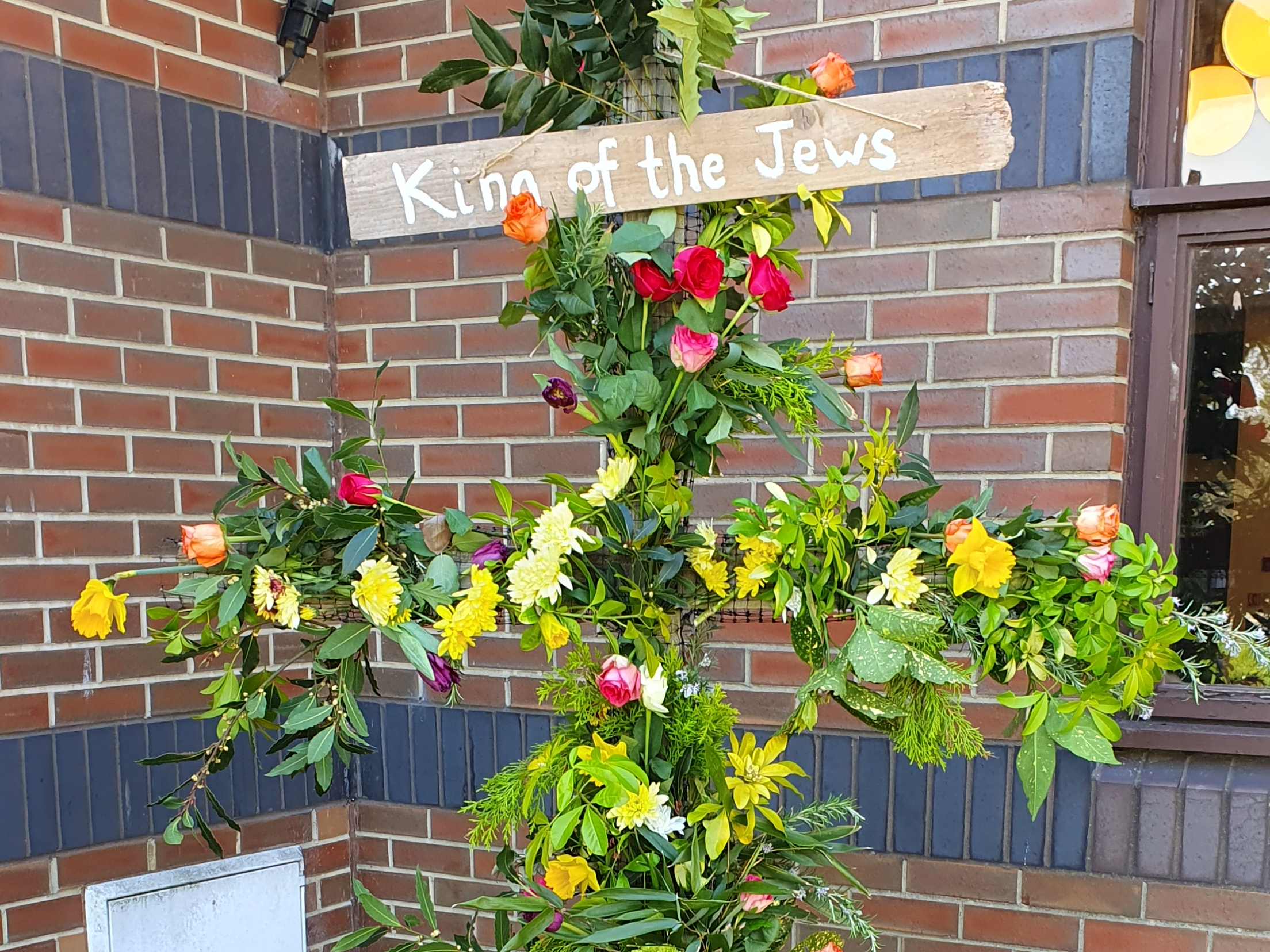 A cross adorned with flowers and greenery. There is a sign at the top saying 'King of the Jews'.