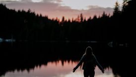 woman standing with hands spread on side facing lake at sundown