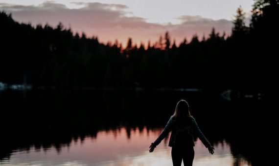 woman standing with hands spread on side facing lake at sundown